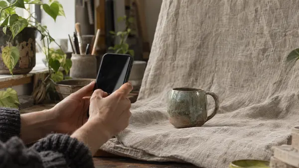 Hands photographing a ceramic mug on a linen backdrop with a phone.